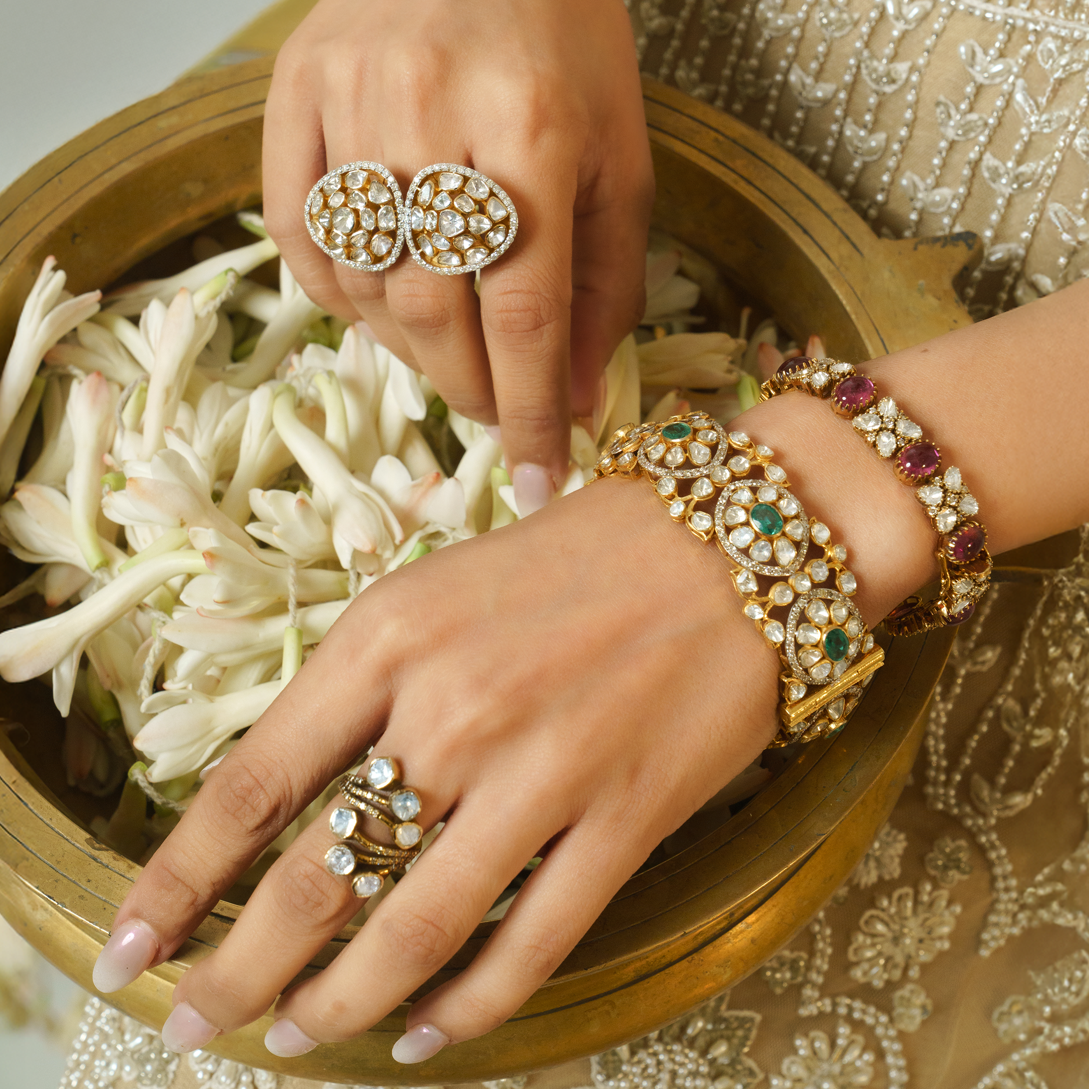 Close-up of hands styled with a wide gold Polki and emerald bangle, a ruby tourmaline and diamond link bracelet, and large statement Polki diamond rings, posed over a brass bowl of white jasmine flowers.