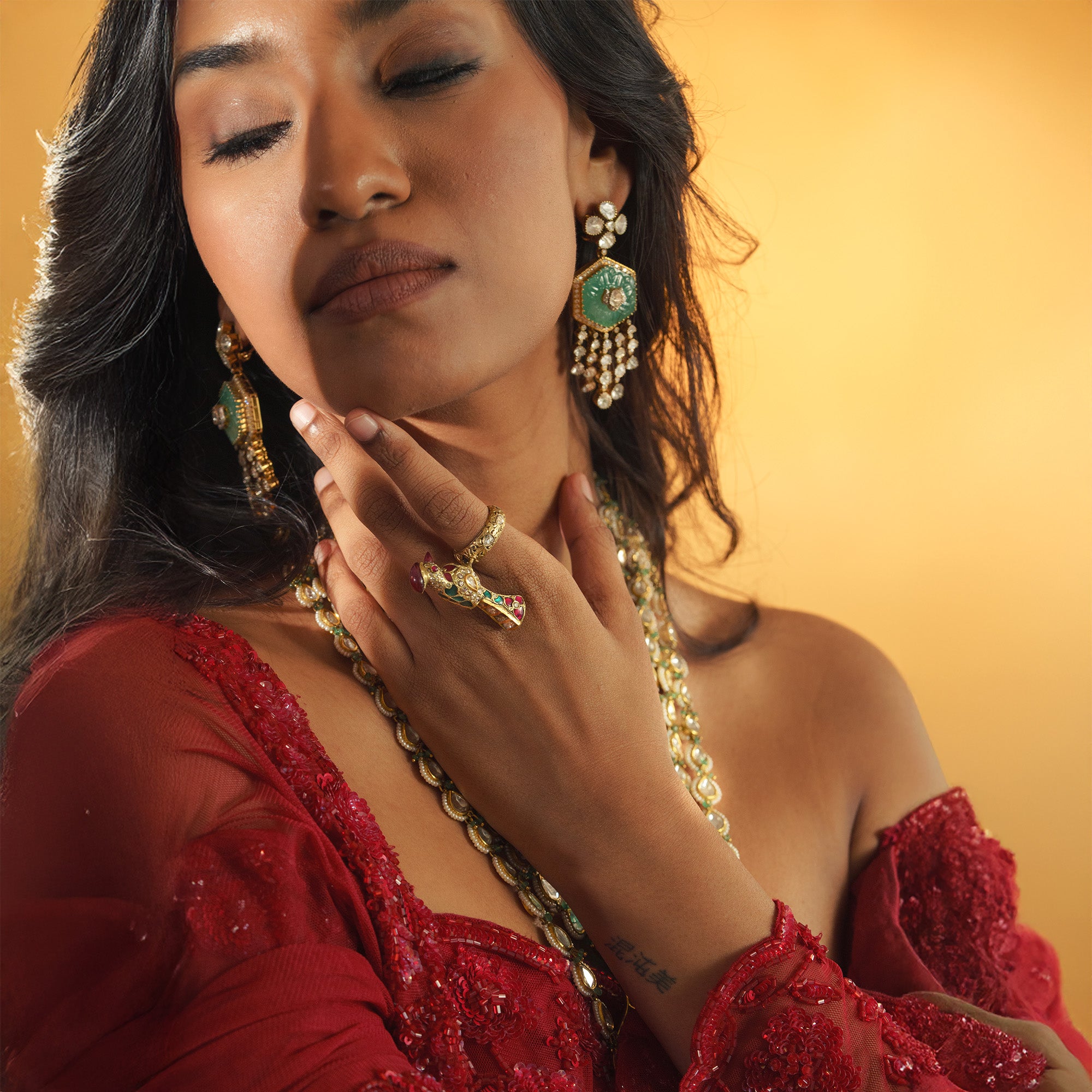 Woman wearing 18kt gold polki emerald ruby jewelry including earrings, necklace, and ring against a warm background in india