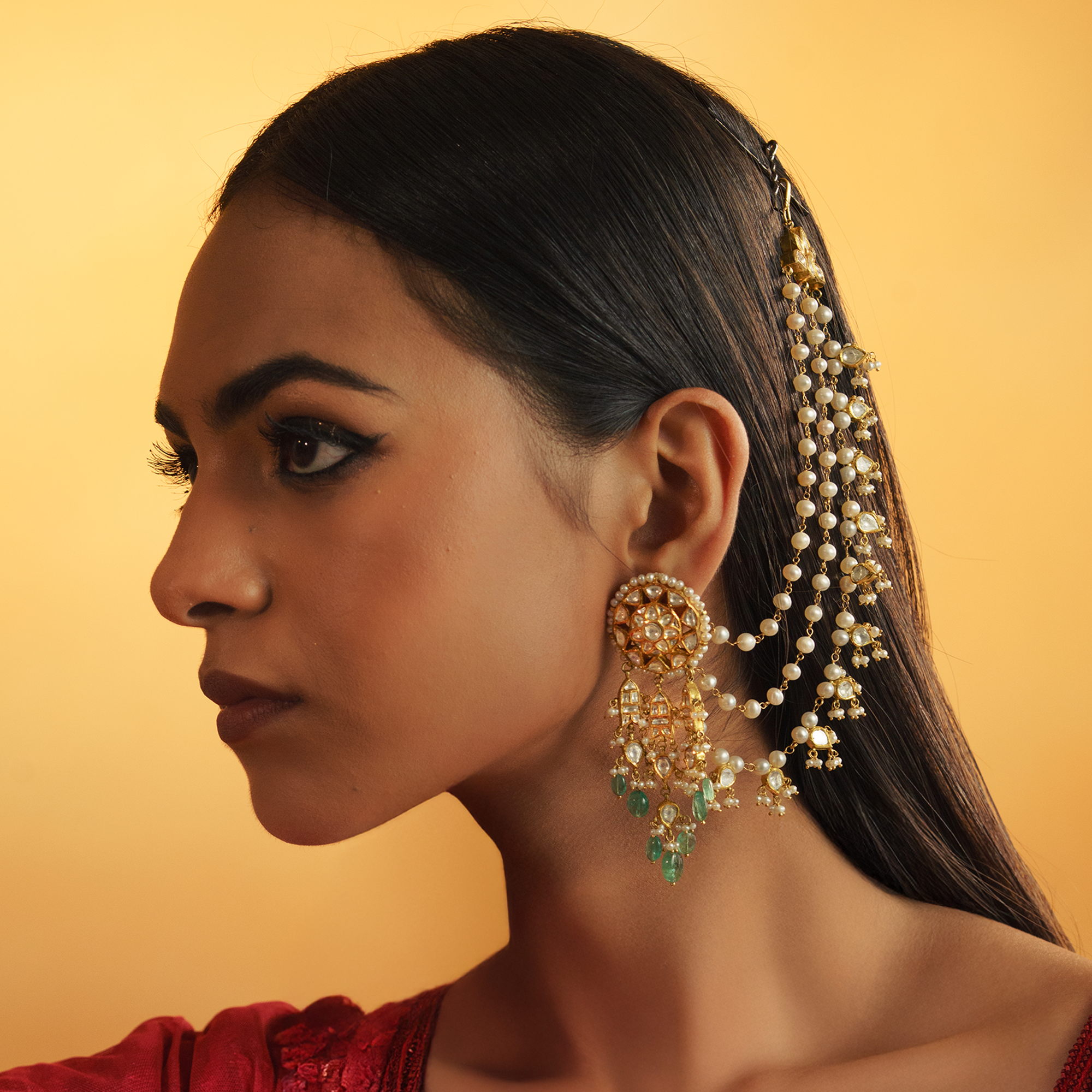 A profile shot of a model wearing ornate polki emerald diamond earrings featuring cascading emerald drops and three-tiered pearl sahara chains pinned into the hair.