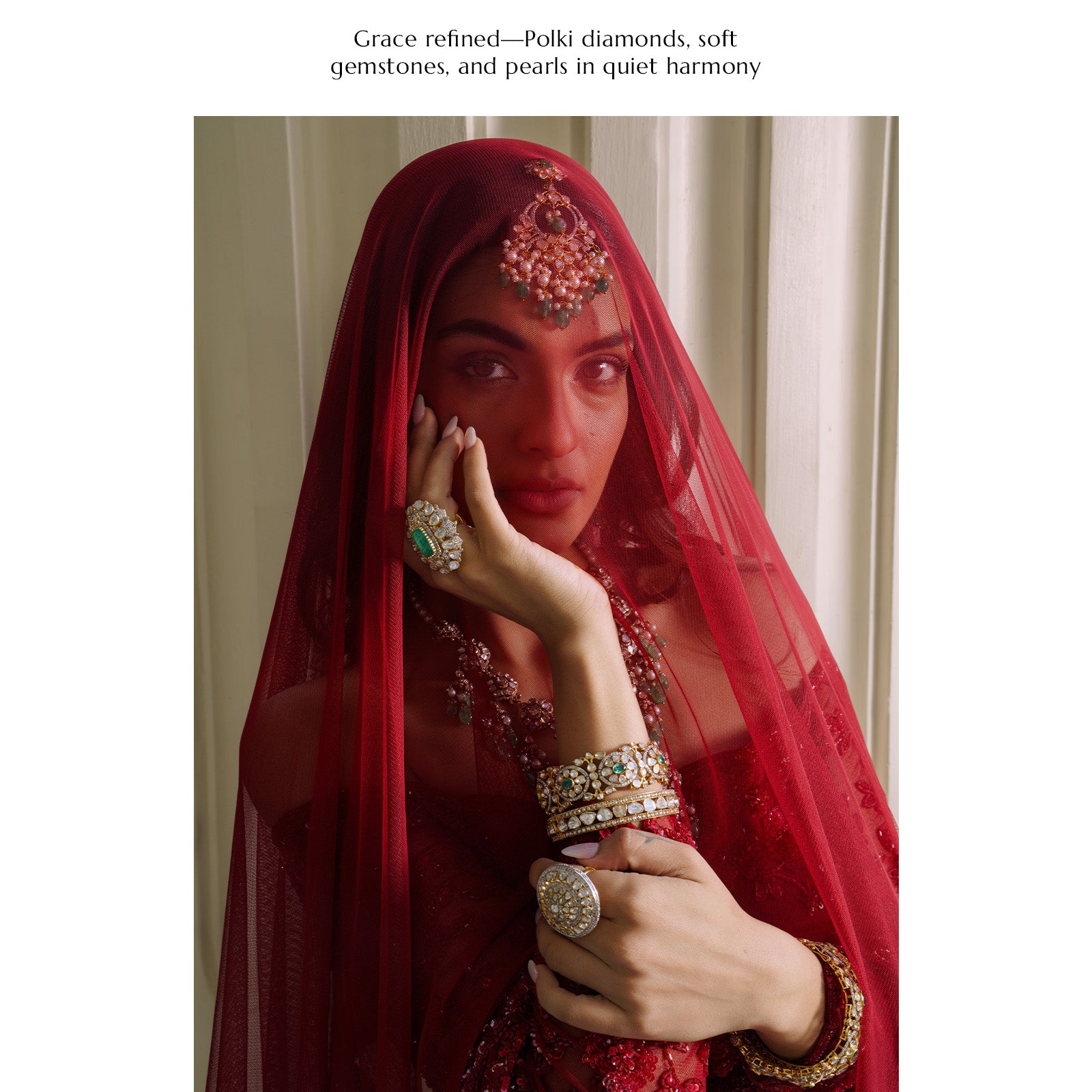 A model wearing a sheer red veil and a full suite of traditional gold jewelry, including an ornate maang tikka with pink stones and emerald drops, a statement emerald and polki cocktail ring, and stacked polki diamond bangles.