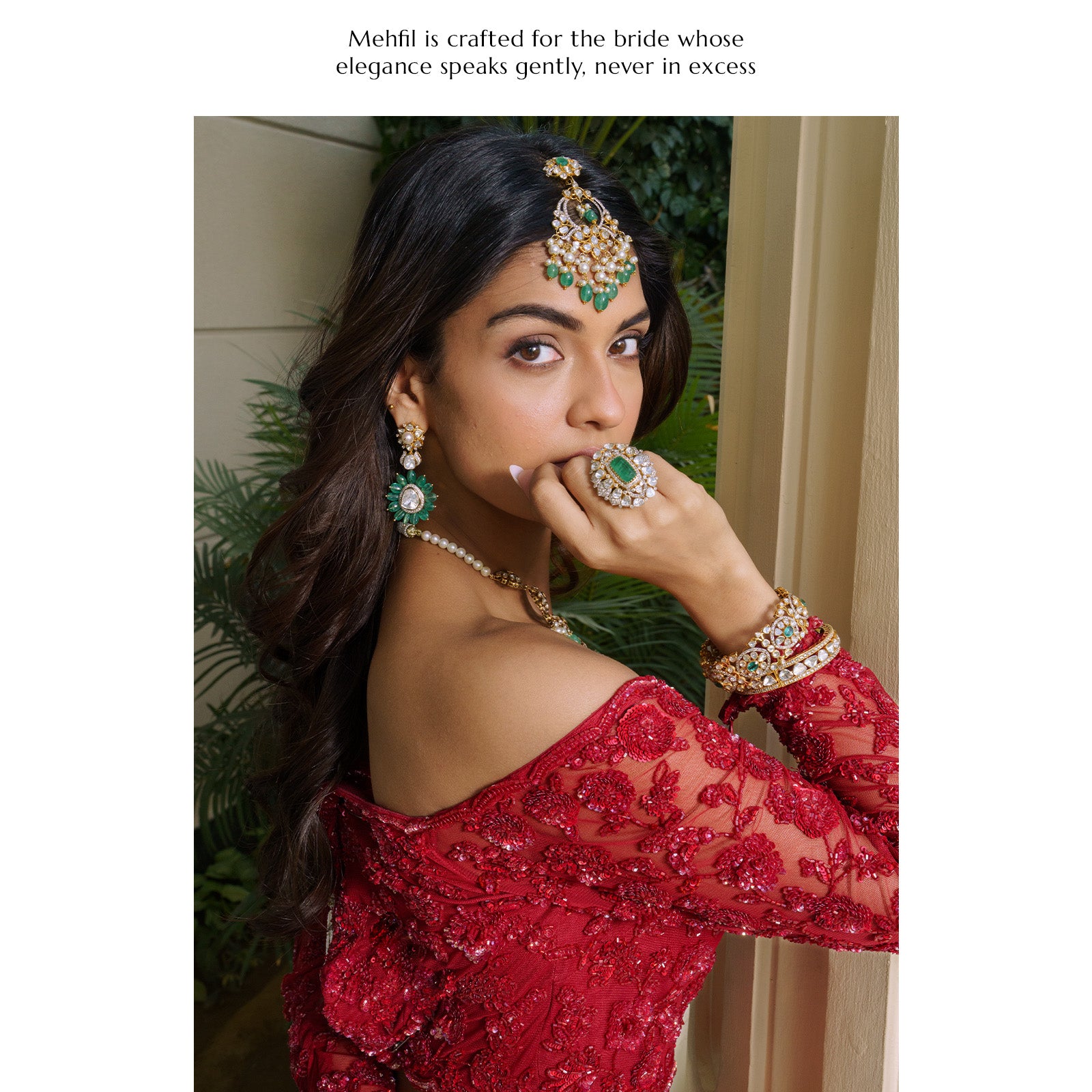 A model wearing an off-the-shoulder red lace ensemble, featuring an ornate emerald and polki maang tikka with a pearl fringe, matching floral-inspired emerald drop earrings, a statement emerald cocktail ring, and stacked polki diamond bangles.