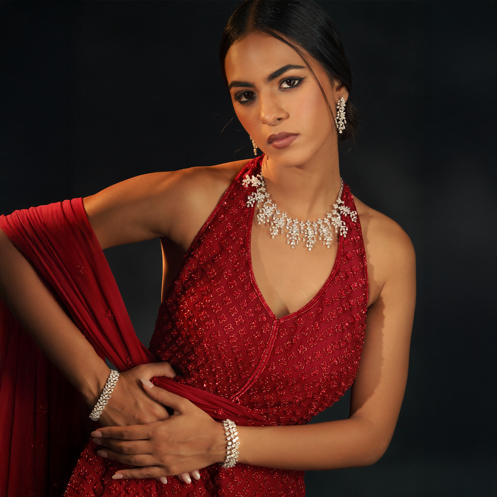 A model in a crimson halter-neck gown wearing a white gold or diamond fringe necklace with delicate, branch-like drops. The look is paired with matching diamond drop earrings and two stacked diamond line bracelets.