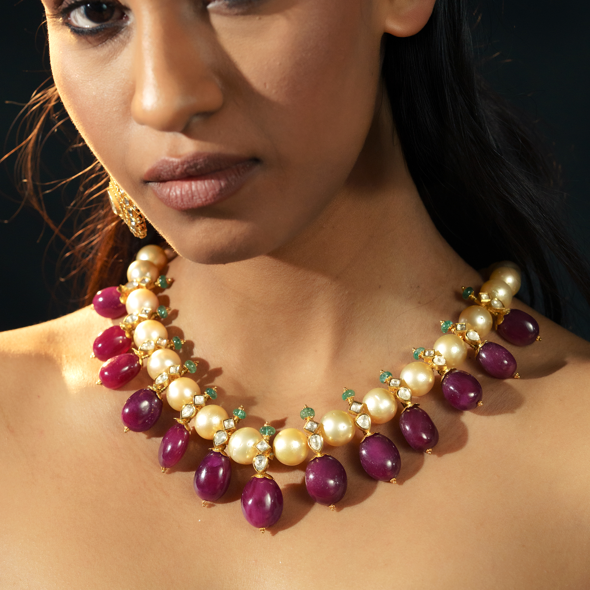 A model wearing a statement gold necklace featuring a row of large, lustrous South Sea pearls accented with tiny emerald beads, finished with a heavy fringe of vibrant ruby drops in india