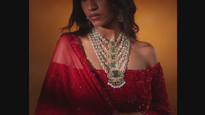 A pair of striking emerald and polki diamond drop earrings featuring large rectangular emerald cabochons with double diamond halos, suspended from triangular polki diamond studs.
Close-up of a hand resting against red floral embroidery, wearing a unique gold bird-motif wrap ring set with rubies and emeralds, paired with a wide gold Polki diamond bangle finished with delicate pearl-strand borders.